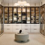 Large boutique closet for her with glass-front cabinets with light tone panels and onyx frames and pulls, soft-close drawers with light colored fronts and onyx hardware, closed cabinets above, all surrounding a center ottoman, and integrated lighting throughout