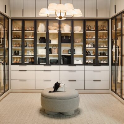Large boutique closet for her with glass-front cabinets with light tone panels and onyx frames and pulls, soft-close drawers with light colored fronts and onyx hardware, closed cabinets above, all surrounding a center ottoman, and integrated lighting throughout