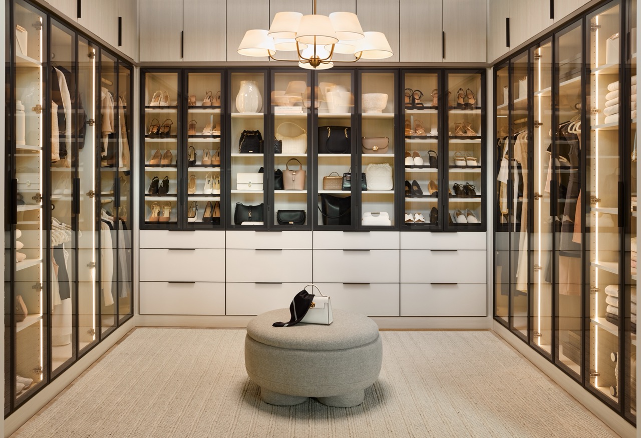 Large boutique closet for her with glass-front cabinets with light tone panels and onyx frames and pulls, soft-close drawers with light colored fronts and onyx hardware, closed cabinets above, all surrounding a center ottoman, and integrated lighting throughout