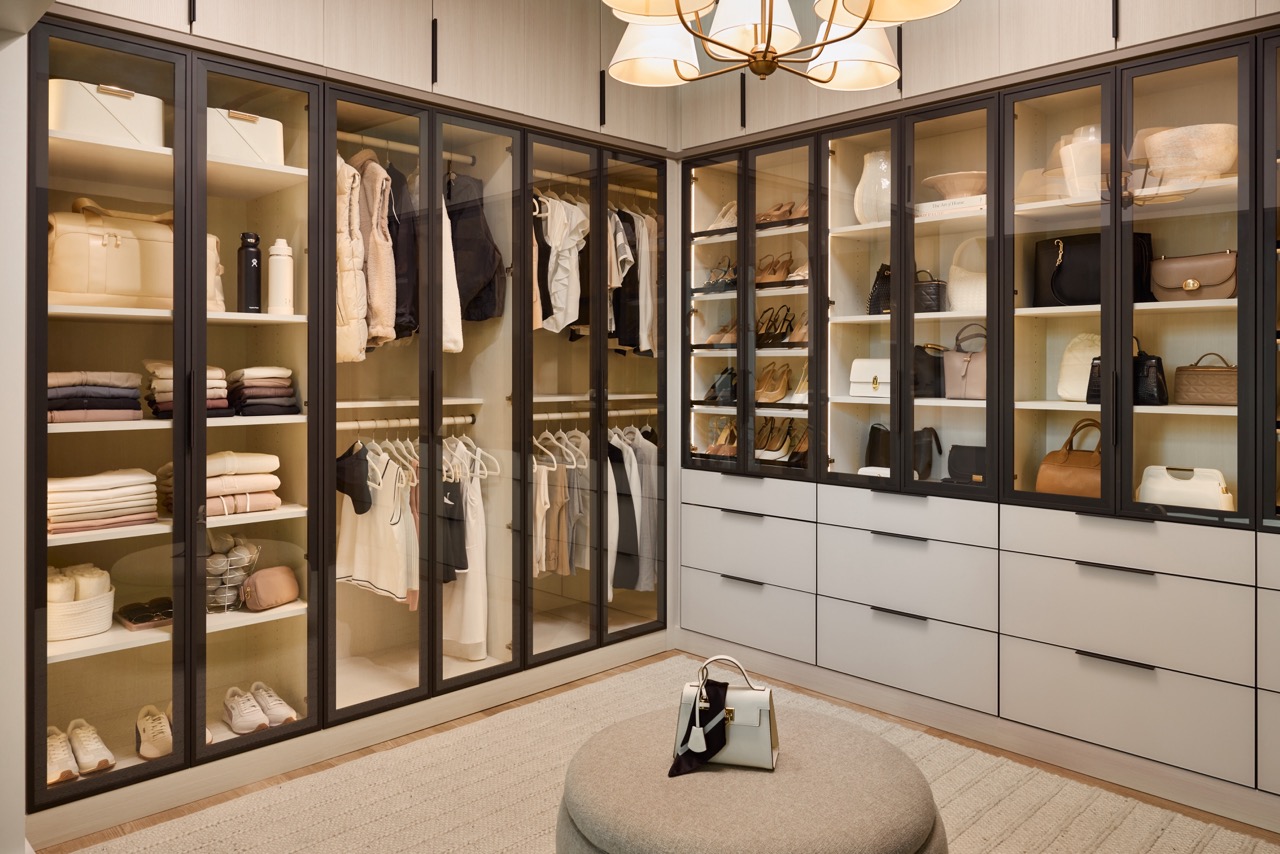 Large boutique closet for her with glass-front cabinets with light tone panels and onyx frames and pulls, soft-close drawers with light colored fronts and onyx hardware, closed cabinets above, and integrated lighting