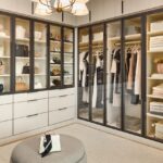 Large boutique closet for her with glass-front cabinets with light tone panels and onyx frames and pulls, soft-close drawers with light colored fronts and onyx hardware, closed cabinets above, and integrated lighting