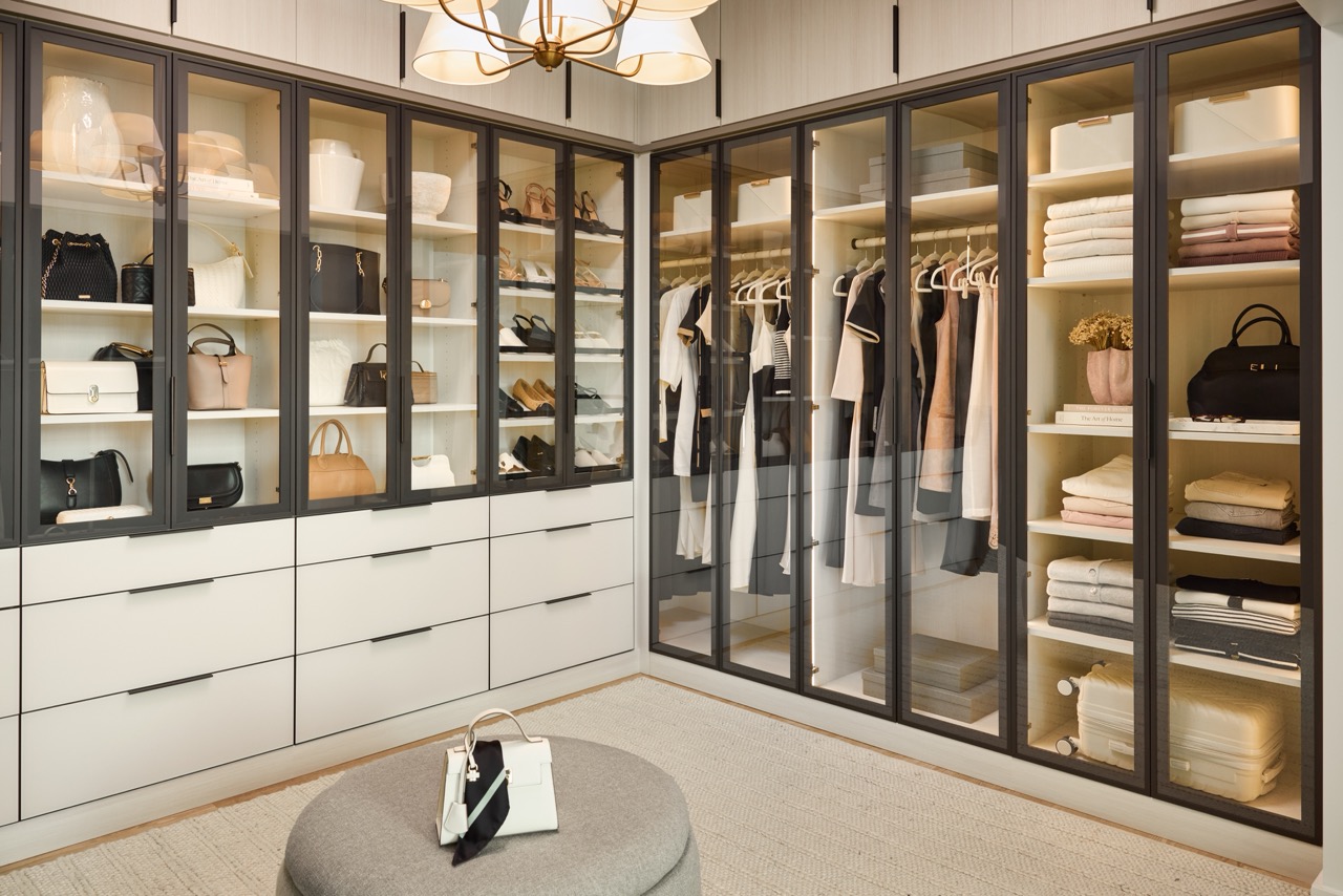 Large boutique closet for her with glass-front cabinets with light tone panels and onyx frames and pulls, soft-close drawers with light colored fronts and onyx hardware, closed cabinets above, and integrated lighting
