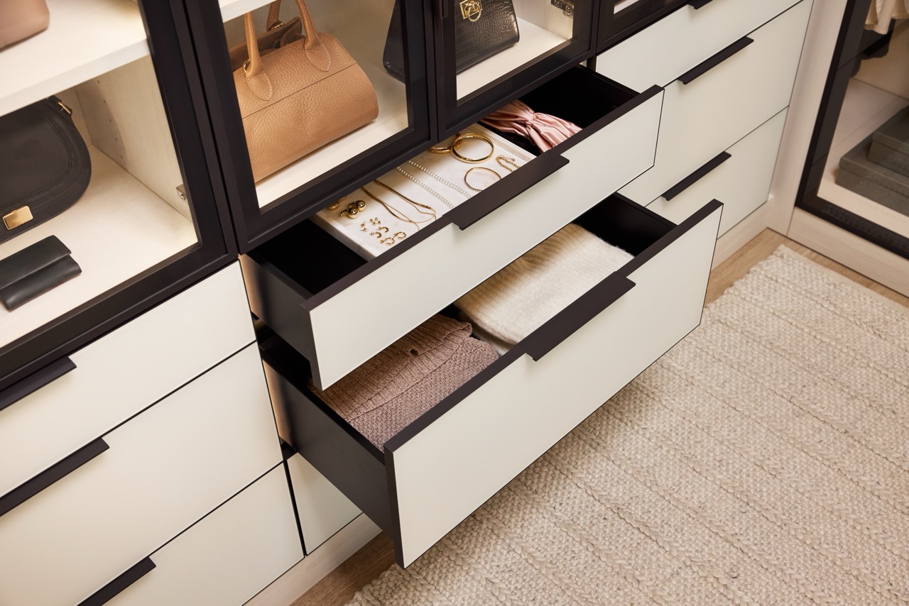 Close up of custom storage in a boutique closet, soft-close drawers with light colored fronts holding jewelry and sweaters, glass-front cabinets with onyx frames holding purses