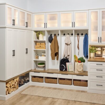 Custom entryway with glass-front cabinets above, and a mix of solid doors, hook storage, open shelves, wicker baskets, and soft-close drawers below holding coats, firewood, backpack, and personal items, in a warm white finish, medium wood tone tops, and matte black hardware