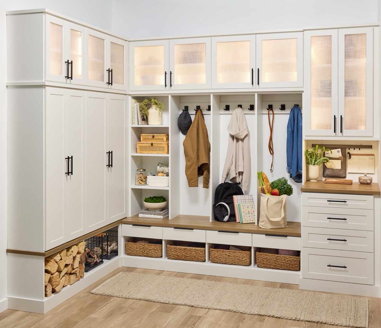 Entryway 1 Custom entryway with glass-front cabinets above, and a mix of solid doors, hook storage, open shelves, wicker baskets, and soft-close drawers below holding coats, firewood, backpack, and personal items, in a warm white finish, medium wood tone tops, and matte black hardware