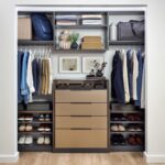 His reach-in closet featuring soft-close drawers with Seared brown fronts, hanging clothes rods, open shelf storage holding shoes, duffle bag, folded clothes, personal items and accessories, in a mix of medium wood and onyx tones, Elusion glass top, and matte black hardware