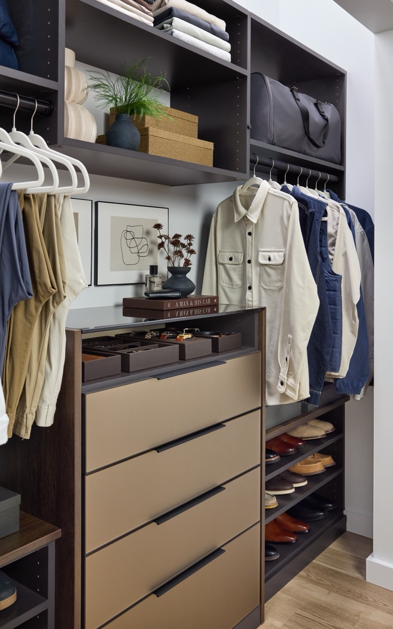 His reach-in closet featuring soft-close drawers with Seared brown fronts, hanging clothes rods, open shelf storage holding shoes, duffle bag, folded clothes, and personal items, in a mix of medium wood and onyx tones, Elusion glass top, and matte black hardware