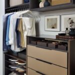His reach-in closet featuring soft-close drawers with Seared brown fronts, hanging clothes rod, open shelf storage holding shoes and personal items, in a mix of medium wood and onyx tones, Elusion glass top, and matte black hardware