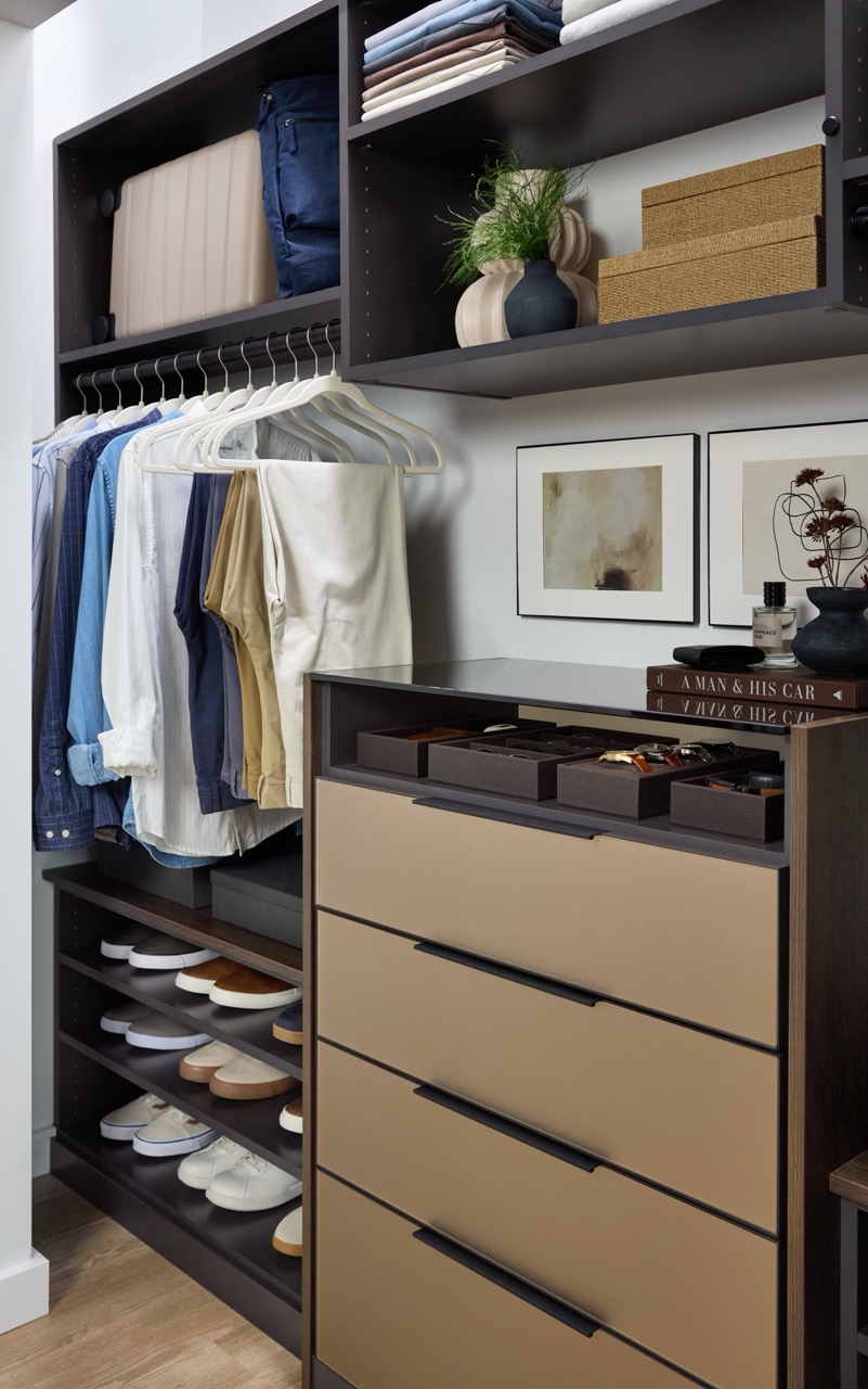 His reach-in closet featuring soft-close drawers with Seared brown fronts, hanging clothes rod, open shelf storage holding shoes and personal items, in a mix of medium wood and onyx tones, Elusion glass top, and matte black hardware