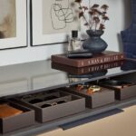 Close up of a man's closet storage solution featuring Elusion glass above an open shelf with leather boxes holding sunglasses, watches, and belts, in medium wood and charcoal tones, Seared brown drawer front, and matte black hardware