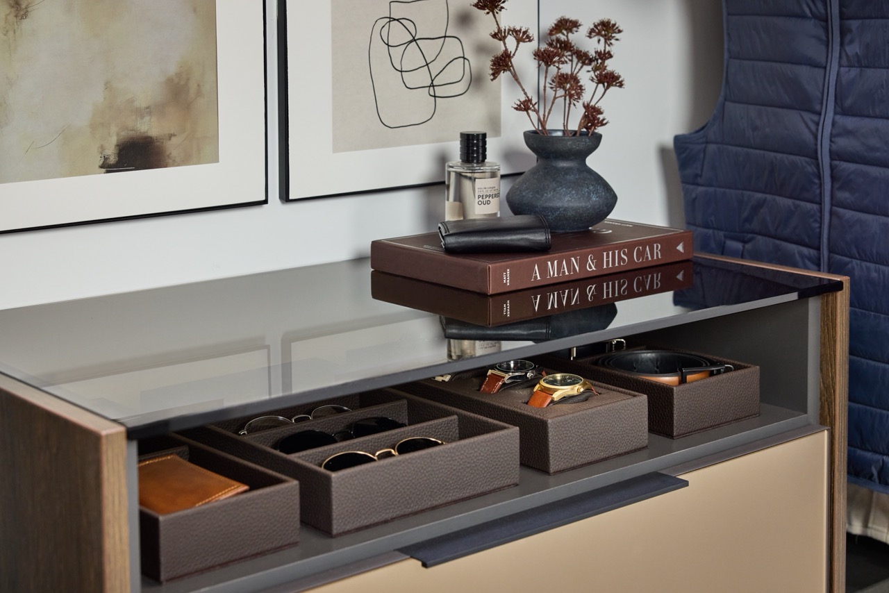 Close up of a man's closet storage solution featuring Elusion glass above an open shelf with leather boxes holding sunglasses, watches, and belts, in medium wood and charcoal tones, Seared brown drawer front, and matte black hardware