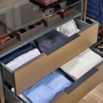 Close up of a man's reach-in closet with soft-close drawers, Elusion glass top, and open shelf storage for personal items in medium wood and charcoal tones, with Seared brown drawer fronts, and matte black hardware
