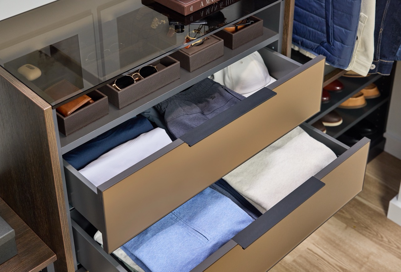 Close up of a man's reach-in closet with soft-close drawers, Elusion glass top, and open shelf storage for personal items in medium wood and charcoal tones, with Seared brown drawer fronts, and matte black hardware