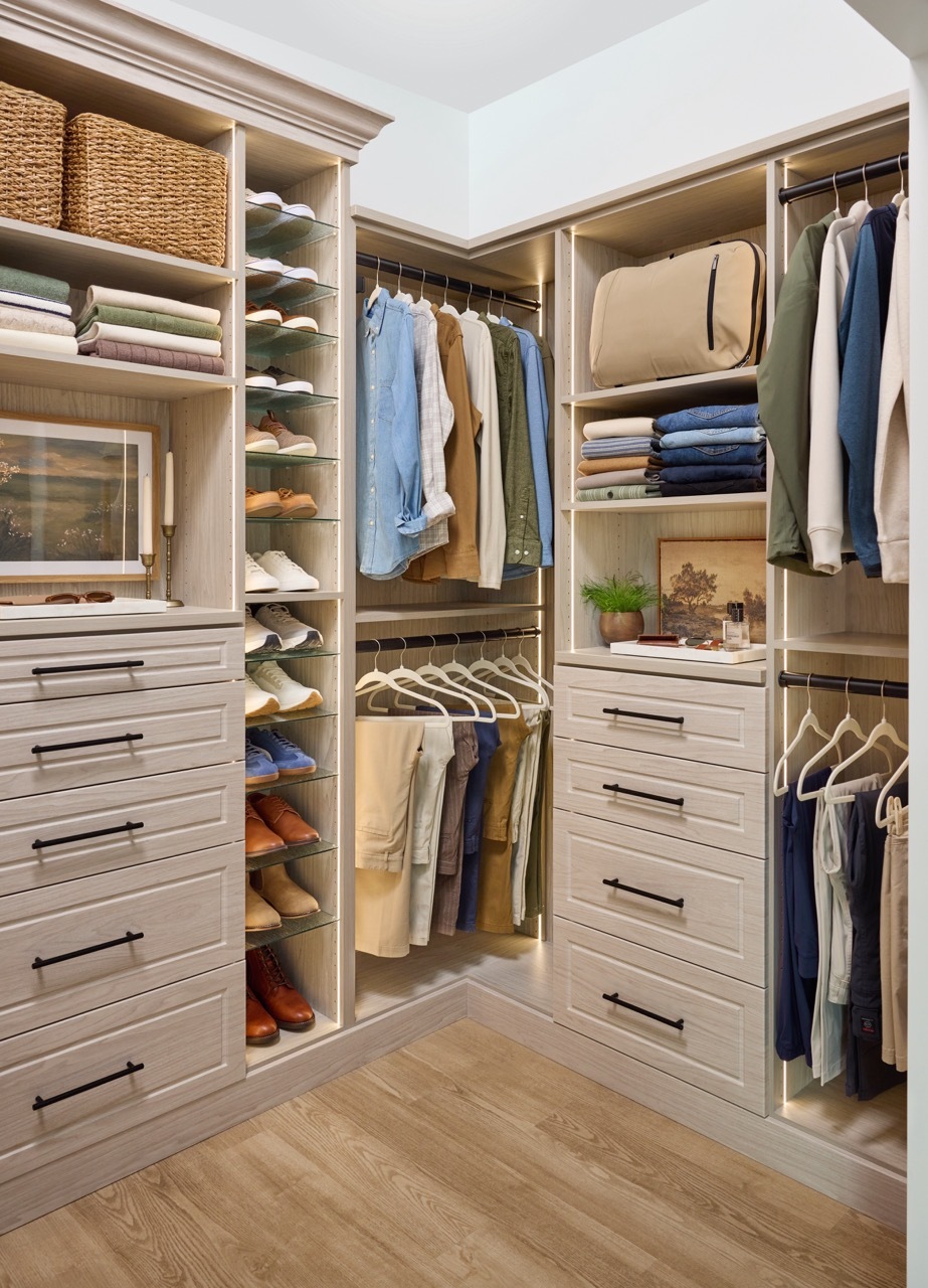 Shared walk-in closet in a light wood-tone finish with custom storage, Shoe Shrine, hanging rods holding a man's shirts and pants, and storage shelves and drawers